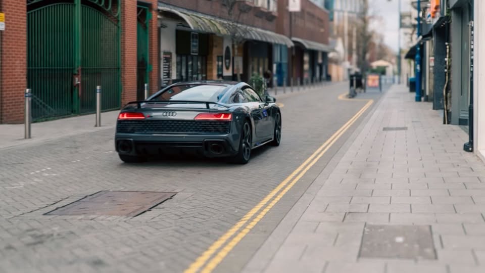Rear view of a dark grey Audi R8 driving down a narrow city street